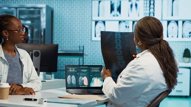 Black patient interacting with a female physician in a medical cabinet, discuss MRI and x ray analysis for personalized care. Medic offers wellness and guidance during scan interpretation. Camera A.