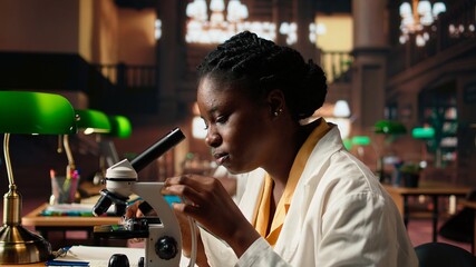 African american scholar studies medical science in a college library, doing research on viruses using a microscope. Girl works on exam preparation for success in a scientific career. Camera A.