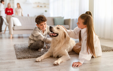 Two cute kids sitting on floor at home, petting golden retriever dog, their parents and granny on background. Multi generation family spending time together in living room