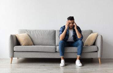 Fototapeta premium Sad young Middle Eastern man suffering from headache or migraine, touching head massaging temples. Stressed guy sitting on couch with painful face expression feeling terrible weakness or depression