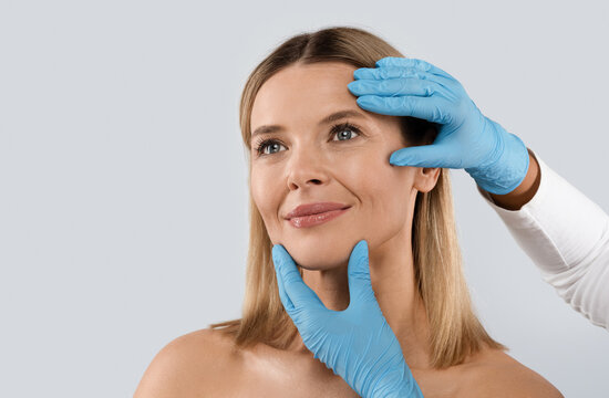 Plastic surgery or face lifting for middle aged woman. Doctor hands wearing blue medical gloves holding head of female patient, checking skin before procedure, isolated on grey background, copy space