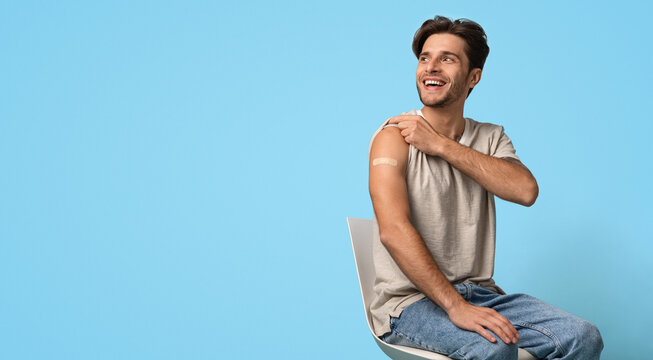 Immunization Campaign. Vaccinated Young Man Showing Arm With Adhesive Bandage After Vaccine Injection And Looking Away, Smiling Guy Sitting On Chair Over Blue Studio Background, Panorama, Copy Space