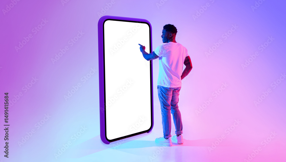 Poster Full length of millennial black guy standing near big cellphone with mockup for your app on screen, interacting with user interface in neon light. Smartphone display template for website or ad