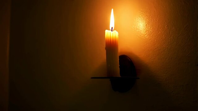 Illuminated wax candle with a flame on decorative metal wall sconce casting a warm glow in a dark room interior at night.