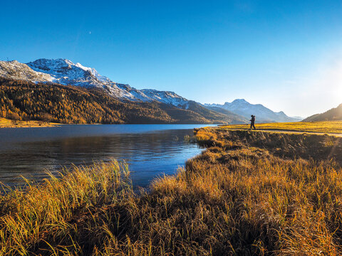 Lone hiker by calm alpine lake with autumn grass and snowy mountains under golden evening sunlight.  Beautiful landscape in the Swiss Alps on a day in autumn.