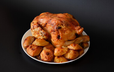 Whole baked chicken and new potatoes on a round ceramic plate on a black background.