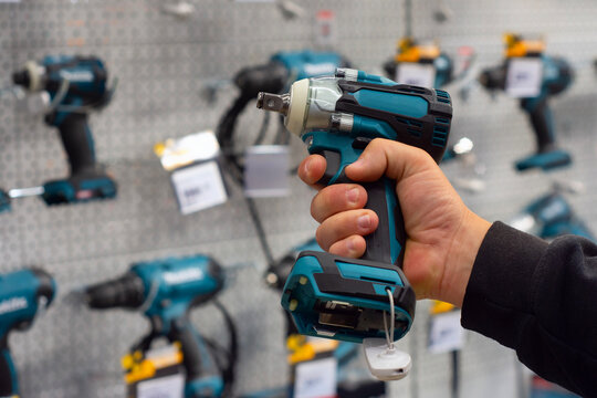 A hand holds a teal power drill in front of a hardware store display with various tools. Concept power tools and construction supplies.
