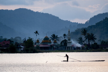 Fisherman on a bamboo raft with traditional Minangkabau houses and hills in West Sumatra,...