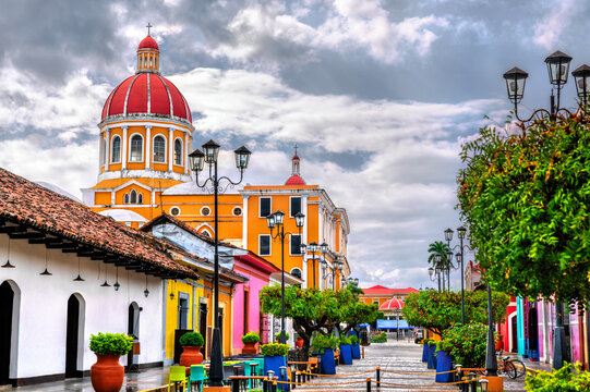 View of Granada's iconic cathedral dome rising behind a vibrant pedestrian street lined with colorful colonial buildings, trees in blue planters, and iron lampposts under a moody sky in Nicaragua.