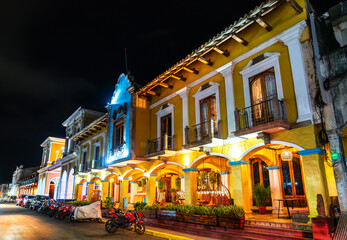 A historic colonial-era building with arched porticoes and balconies glows under streetlights at the Central Park of Granada, Nicaragua. The nighttime setting highlights the rich yellow facade and