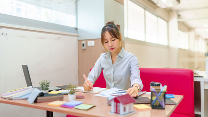 Asian Young Interior Designer Woman Reviewing Blueprints at Desk with Laptop Paint Swatches and Miniature House Model Inspiring Creative Design
