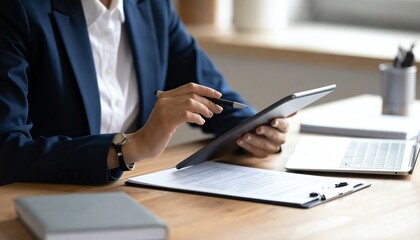 A person in a suit uses a tablet while seated at a desk, surrounded by documents and a laptop, in a professional office setting