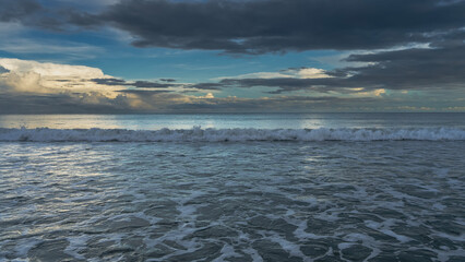 Fototapeta premium Early morning. The clouds in the blue sky are illuminated with golden light. The ocean waves roll towards the shore, foam and spread. Glare, reflection on the surface of the sea. Malaysia. Borneo.