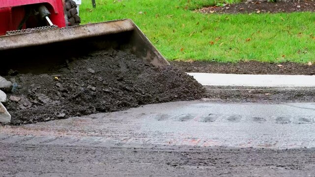  Compact loader , skid steer with the front bucket scooping up asphalt or dirt from a road surface during a paving or road repair project.