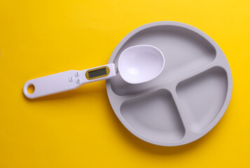 Silicone plate with dividers for feeding baby and spoon scales on yellow background. Top view