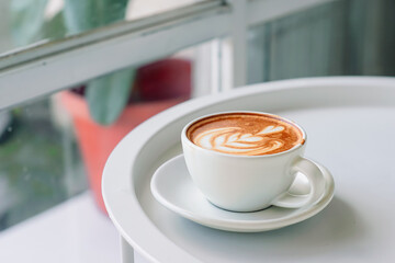 A cup of cappuccino with latte art on a white table near a window.