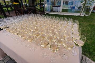 A large number of wine glasses filled with white wine arranged on a table outdoors, ready for an...