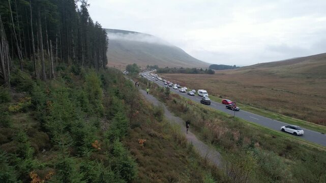 People walking on pathway leading parallel to road. Row of cars parking on grass along road. Static shot. Brecon Beacons national Park, Wales, UK
