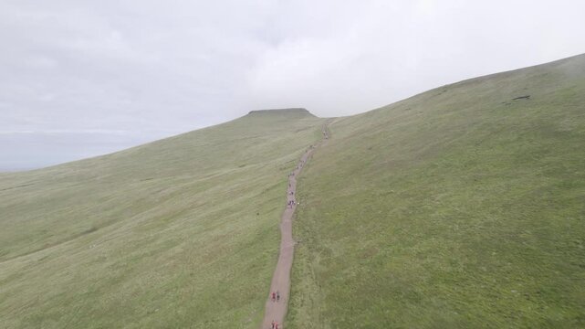 Busy tourist path in green grassy slope. Forwards fly above people hiking in mountains on cloudy day. Brecon Beacons national Park, Wales, UK