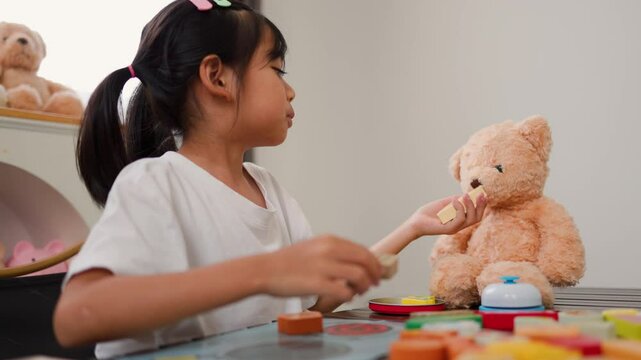 Asian girl engaging in pretend play with teddy bear and toy food at home. Encouraging creativity, social skills, and early childhood development through imaginative learning activities.