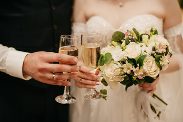 The bride and groom hold glasses of champagne and a wedding bouquet of large white roses in their hands during the wedding walk.