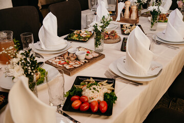 A variety of food is on a table covered with a white tablecloth, empty plates with cloth napkins on them in the shape of a cone, flowers in vases stand between the dishes