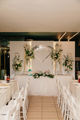 banquet hall with tables set for a wedding celebration with white chairs and a table in the center of the hall against the background of a white screen decorated with fresh flowers