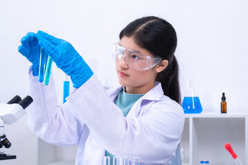 A young Asian female scientist in lab coat and gloves holds a glass pipette carefully in a science...