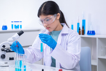 A young Asian female scientist in lab coat and gloves holds a glass pipette carefully in a science...