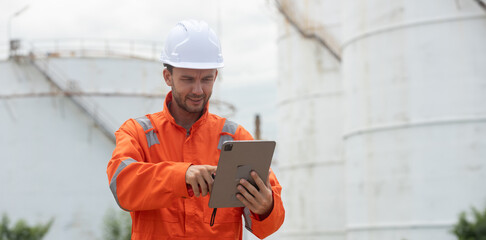 Worker in safety gear using tablet near industrial storage oil tanks refining petrochemical...