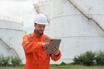 Worker in safety gear using tablet near industrial storage oil tanks refining petrochemical...