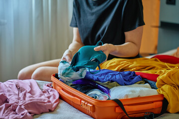 Woman folds colorful clothes into orange suitcase in bedroom. Concept of vacation preparation,...