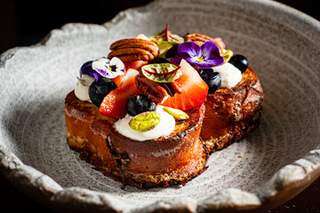A lovely brioche dessert topped with fruits, nuts, and flowers on a rustic plate