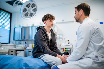 Depressed teenage patient sitting on hospital bed, talking with caring doctor.