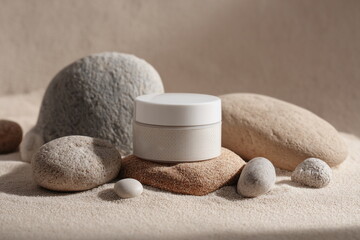 A white skincare jar sits on beach sand among smooth stones, symbolizing purity, natural care and the calm energy of mineral-based beauty rituals.

