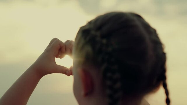 Hands shape heart. Little girl make sign love hands sunset. Small kid draw heart hands against sky. Sign heart symbol love, feeling. Silhouette of heart hands in sun close-up. Sunlight between fingers