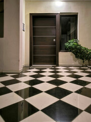 Modern entrance dark wooden door with horizontal metal strips, window, and potted plant. Floor is adorned with black and white checkered tile pattern, and the space is illuminated by a ceiling light.