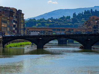 Florence, Italy. Trinity bridge crosses the Arno river featuring three stone arches, on calm waters with reflections. Links bustling city life with serene natural beauty.