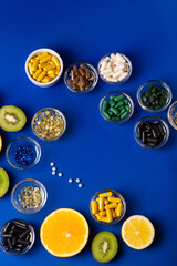 Many vitamins and minerals, dietary supplements in small glass bowls, orange, kiwi, lime and lemon as sources of natural vitaminsa nd minerals. Vitamins and fruits from above on a blue background
