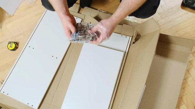 Handyman takes golden hinges out of plastic bag over furniture components set on floor closeup. Worker prepares fastening elements of cabinet door