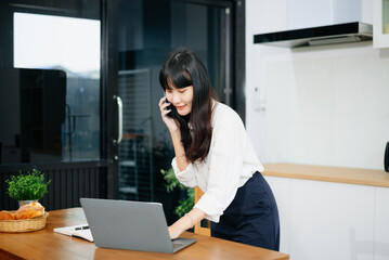 Obraz premium Asian woman working from home while talking on phone in kitchen. Perfect for remote work