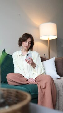 happy girl woman sitting at home on the couch looking at her phone and scrolling through her news feed. cozy living room interior