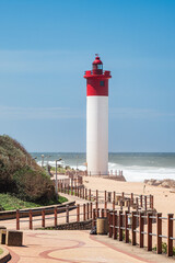 A striking red and white lighthouse stands tall on a sunny day along the Umhlanga beachfront promenade in KwaZulu-Natal, South Africa. The clear blue sky, coastal walkway, and rolling ocean waves. © Brad