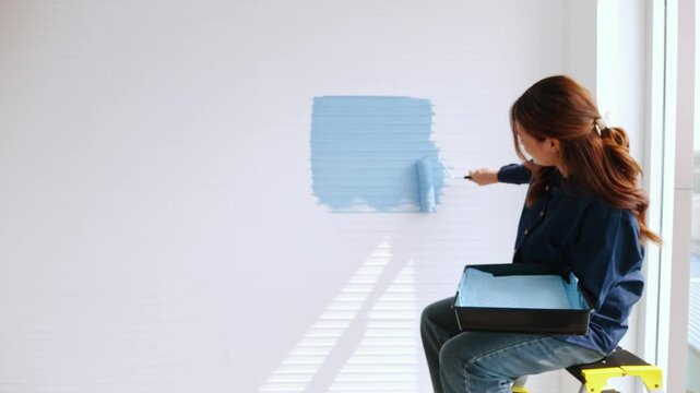 Young woman painting a wall blue with a roller while seated on a step stool, holding a paint tray&mdash;capturing a DIY home improvement scene filled with creativity and modern interior design.