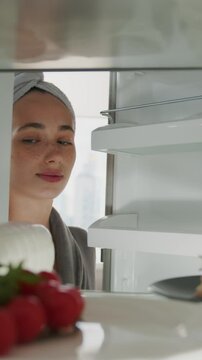 Beautiful caucasian woman in robe opens refrigerator full of healthy food and takes a cake. POV from inside the frige.