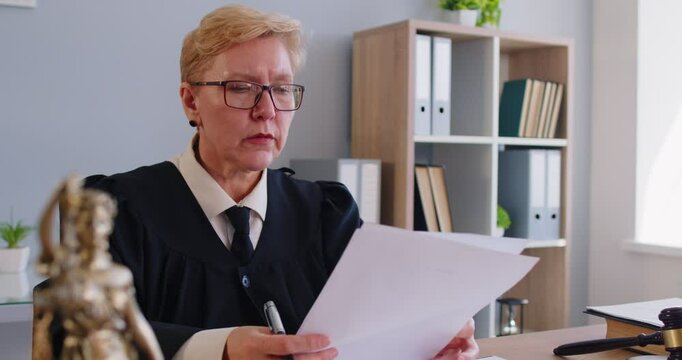 Seious woman judge looking through judiciary papers, dressed in robe gown uniform and glasses, posing at desk in her cabinet, doing investigation, studies case. Law and justice concept