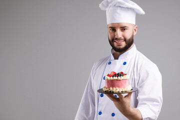 Happy confectioner in uniform holding delicious cake with berries on light grey background