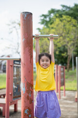 Fototapeta premium Happy Asian girl demonstrating strength and joy while hanging from a playground bar during sunny day in a vibrant outdoor park setting with green trees