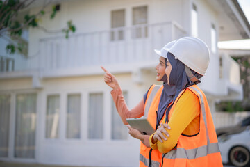 Confident Muslim women architects working at a building site. Wearing orange safety vests and helmets, they point and smile while holding construction plans and digital tablet.