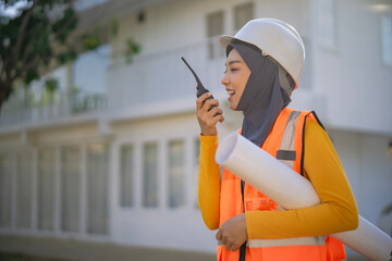 Muslim female construction manager holds blueprints and a two-way radio while inspecting a housing site. She represents modern engineering and leadership in field work...